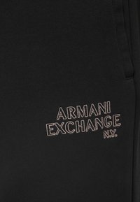 Tecido preto com texto bordado em bege "ARMANI EXCHANGE N.Y." no lado direito, apresentando uma costura vertical e a borda de um bolso parcial.