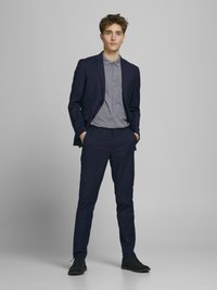 Marineblaues Anzug mit taillierter Jacke, passenden Hosen und einem grauen gemusterten Poloshirt. Strukturierten Stoff, Button-Down-Kragen und schwarze Schuhe.