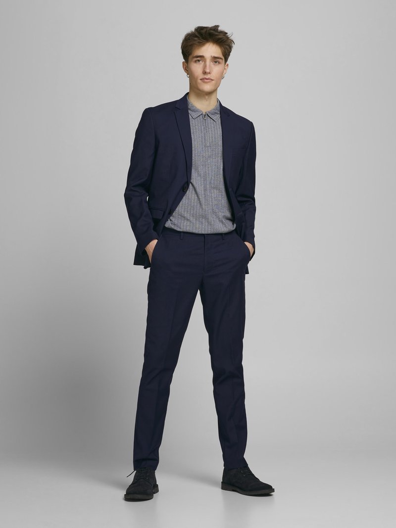 Marineblaues Anzug mit taillierter Jacke, passenden Hosen und einem grauen gemusterten Poloshirt. Strukturierten Stoff, Button-Down-Kragen und schwarze Schuhe.