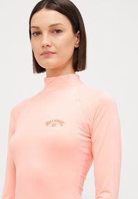 Rash guard a maniche lunghe di colore rosa chiaro, realizzato in tessuto elasticizzato. Presenta un collo alto e il logo Billabong in una tonalità arancione a contrasto.