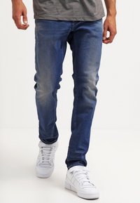 Jean en denim bleu avec une coupe slim, des accents décolorés et une texture lisse. Associés à des baskets blanches avec des détails perforés.