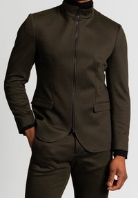 Blazer su misura verde oliva con colletto alto, zip frontale e due tasche laterali. Realizzato in un tessuto liscio e strutturato con un design aderente.