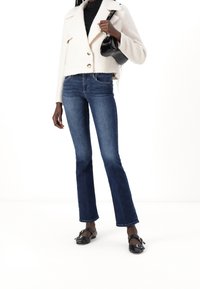Chaqueta blanca texturizada y recortada con grandes botones, combinada con unos jeans acampanados de color azul oscuro. Bolso negro y zapatos planos negros con detalle de hebilla.