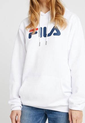 Sudadera con capucha blanca de FILA con logo rojo y azul, llevada por una persona de cabello largo y rubio, combinada con jeans azules sobre un fondo liso.