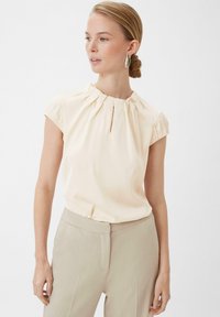 Light beige shirt with a gathered neckline and short sleeves, paired with light beige trousers. Smooth fabric with slight sheen, minimal detailing.