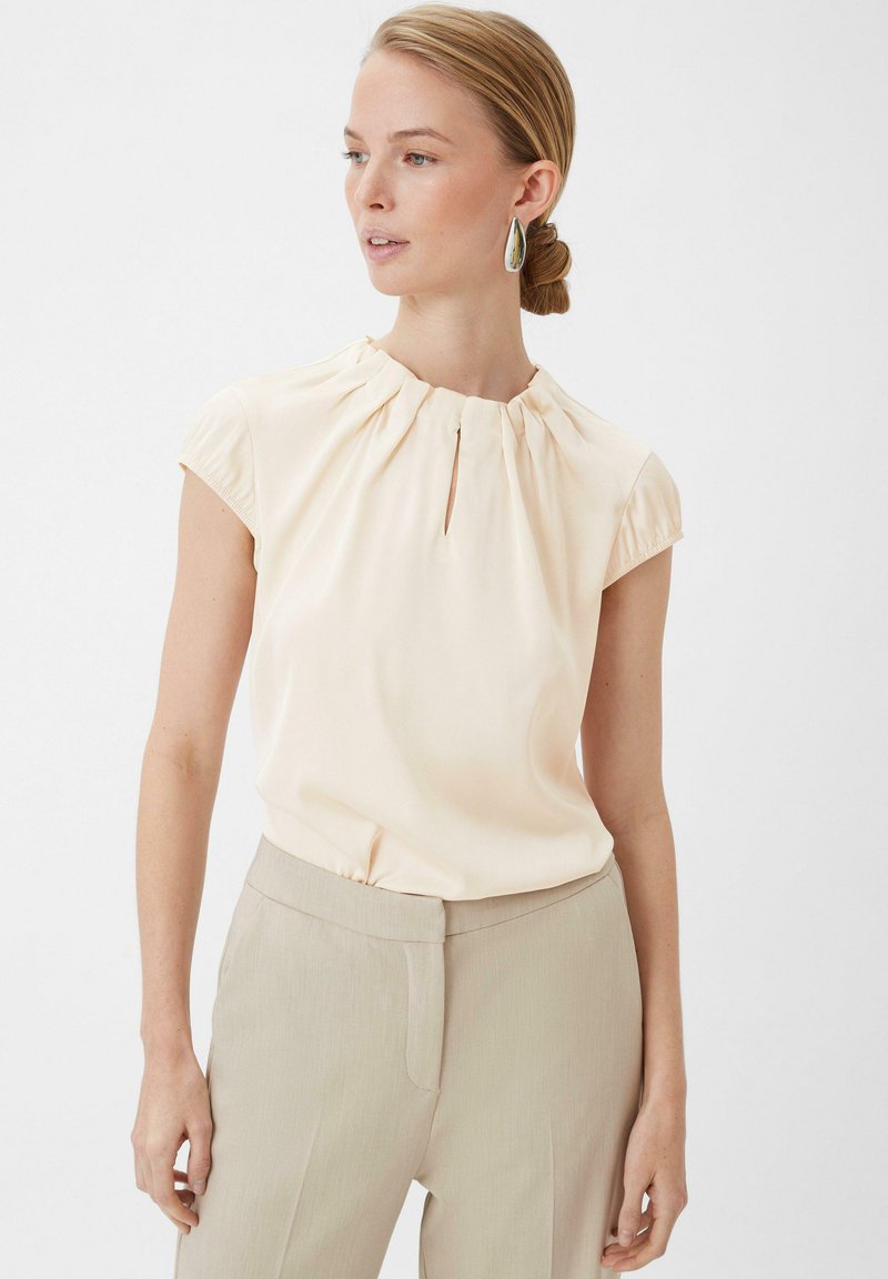 Light beige shirt with a gathered neckline and short sleeves, paired with light beige trousers. Smooth fabric with slight sheen, minimal detailing.
