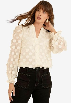 Blusa bianca con pois, scollatura a V, maniche a volant e chiusura con bottoni. Realizzata in tessuto leggero e trasparente con un aspetto strutturato.