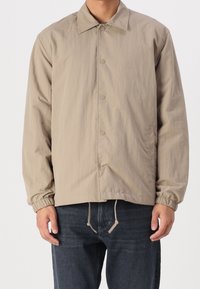 Beżowa lekka kurtka z przodem na guziki, kołnierzem rozłożystym, elastycznymi mankietami oraz regulowanym dołem, wykonana z gładkiej, teksturowanej tkaniny.