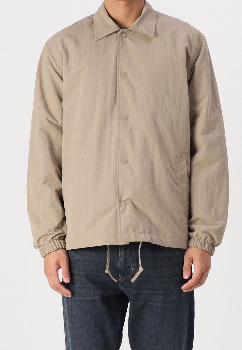 Beżowa lekka kurtka z przodem na guziki, kołnierzem rozłożystym, elastycznymi mankietami oraz regulowanym dołem, wykonana z gładkiej, teksturowanej tkaniny.
