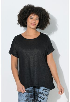 Vrouw met krullend haar die gouden oorringen draagt, een losse zwarte top met korte mouwen en blauwe tie-dye broek met zwarte tekst.