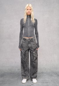 Tailliertes, grau gestreiftes, geripptes Zip-Top und lockere graue Cargo-Hosen mit Taschen, kombiniert mit weißen Sneakers. Der Hintergrund ist minimalistisch grau.