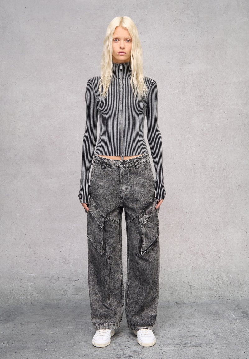 Tailliertes, grau gestreiftes, geripptes Zip-Top und lockere graue Cargo-Hosen mit Taschen, kombiniert mit weißen Sneakers. Der Hintergrund ist minimalistisch grau.