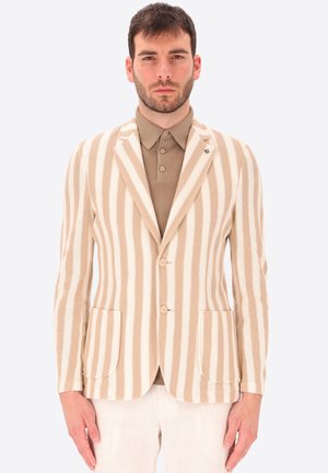 Uomo con blazer beige e bianco a righe verticali sopra una polo taupe con bottoni e pantaloni chiari, in piedi davanti a uno sfondo neutro.