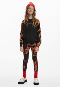 Felpa nera con scritta grafica rossa, abbinata a leggings con motivo a vortice vivace in rosso e giallo. Berretto lavorato a maglia rosso indossato sulla testa.