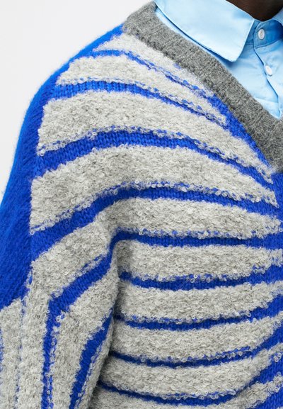 Φλυνωτό πουλόβερ με γκρι και μπλε ρίγες σε διάφορα πλάτη, με σχέδιο V-λαιμού και μαλακή υφή. Ανοιχτό μπλε πουκάμισο από κάτω.