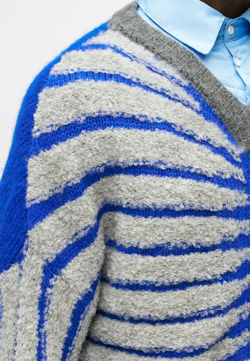 Jersey de punto difuso con rayas grises y azules de varios anchos, con un diseño de escote en V y una textura suave. Camisa de color azul claro debajo.