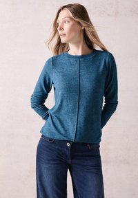 Pull à manches longues en teal avec un col rond, en tricot texturé et un détail de couture centrale. Associé à un jean bleu foncé à jambes larges.