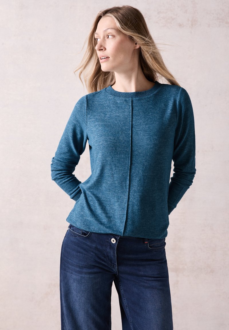 Pull à manches longues en teal avec un col rond, en tricot texturé et un détail de couture centrale. Associé à un jean bleu foncé à jambes larges.