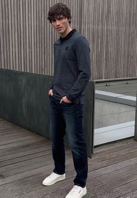 Jonge man staand op een houten dek, handen in de zakken, draagt een donker poloshirt met lange mouwen, donkere spijkerbroek en witte sneakers tegen een grijs en houten paneel achtergrond.