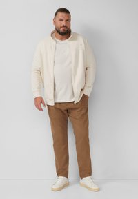 Helle beige Zip-up Jacke über einem weißen Shirt, kombiniert mit braunen Hosen und weißen Sneakers. Schlichtes Design mit minimalen Details.