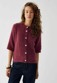 Bordeaux kortærmet cardigan lavet af blødt, fuzzy stof, med hvide knapper foran og en rund halsudskæring.