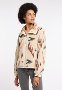 Fleecejacke mit beigem Grundton und mehrfarbigen Zickzackmustern in Braun, Grau und Rostorange; ausgestattet mit einem Reißverschluss und hohem Kragen.