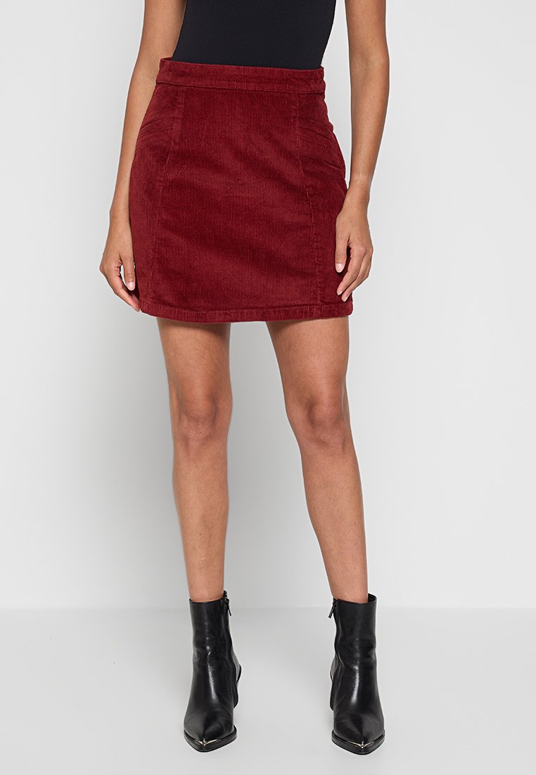 Vrouw in een rode corduroy mini rok, zwart mouwloos topje en zwarte enkellaarsjes die tegen een effen lichte achtergrond staat.