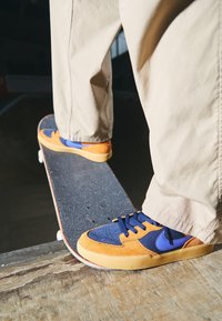 Sneaker med orange mocka och marinblått tyg, med blå detaljer, texturerad tåbox, svarta snören och en tjocktan gummisula på en skateboard.