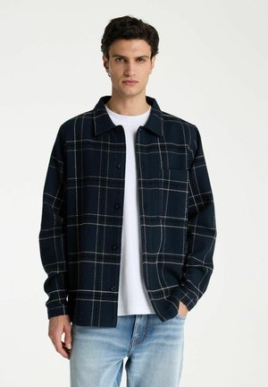 Junger Mann, der ein marineblau kariertes Hemd mit Knöpfen über einem weißen T-Shirt und hellblauen Jeans trägt und vor einem schlichten weißen Hintergrund steht.