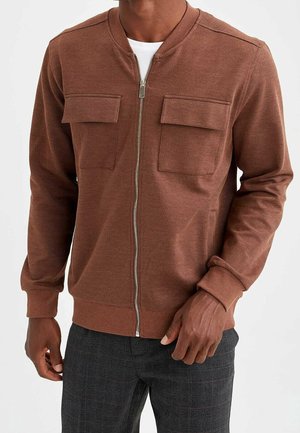 Mann trägt eine braune Jacke mit Reißverschluss, zwei Brusttaschen und dunkle karierte Hosen, Hände locker seitlich.