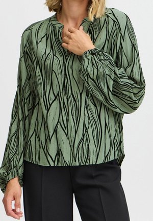Vrouw die een groene blouse met zwart abstract bladerenpatroon en zwarte broek draagt, met haar hand de kraag aanrakend, tegen een effen achtergrond.