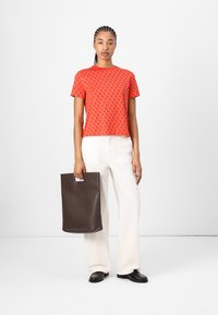 Red cropped t-shirt with tonal crescent pattern, paired with wide cream trousers. Holding a brown leather tote with a cut-out handle.