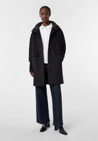Cappotto nero con colletto alto, chiusura a bottoni e tasche laterali, abbinato a una camicia bianca e jeans scuri, caratterizzato da una vestibilità comoda e una texture liscia.