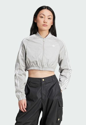 Light gray cropped jacket with a high collar, zip closure, and white stripes on sleeves. Paired with dark cargo pants featuring a gold button.