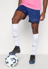 Αθλητικές κοντές φόρμες Nike σε μπλε χρώμα με μαύρες λεπτομέρειες, συνδυασμένες με άσπρες κάλτσες ως το γόνατο που φέρουν λογότυπο, και μαύρα ποδοσφαιρικά παπούτσια πάνω σε μια σχεδιασμένη μπάλα.