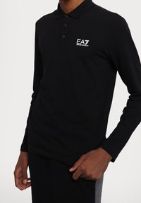 Černá polo košile s dlouhým rukávem vyrobená z bavlny, s límcem na tři knoflíky a bílým logem EA7 Emporio Armani na hrudi.