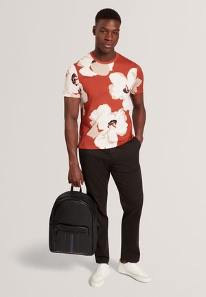 Rotes und weißes Blumen-T-Shirt kombiniert mit schwarzen Hosen und weißen Turnschuhen; hält einen schwarzen Rucksack mit vertikalen Streifen und Reißverschlusstasche.