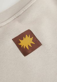 A bézs pulóver anyaga bordázott nyakkal rendelkezik. Egy barna négyzet alakú folt sárga napsugár mintát mutat, hangsúlyozva a textúrát és a színkontrasztot.