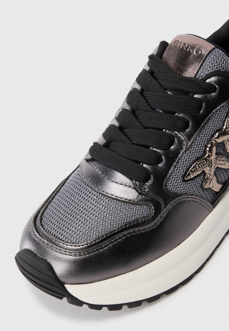 Schwarzer und metallic grauer Sneaker mit Obermaterial aus Mesh und Leder, runder Zehenpartie, schwarzen Schnürsenkeln und Logo-Akzent an der Seite. Weiße Zwischensohle und strukturierte Außensohle.