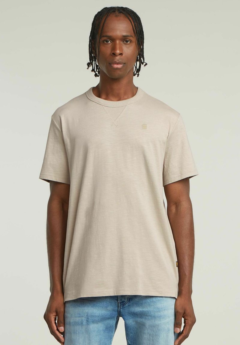 T-shirt beige a maniche corte con scollatura a costine, morbido al tatto e con un piccolo logo sul petto. Abbinato a jeans in denim azzurri.