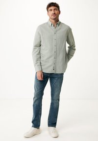 Světle zelená košile na knoflíky s límečkem, kombinovaná s modrými džíny a bílými teniskami. Košile má texturovanou tkaninu a dlouhé rukávy.