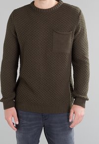 Maglione lavorato a maglia verde oliva con un motivo testurizzato, scollo rotondo e taschino sul lato sinistro del petto. Polsini e orlo a coste.
