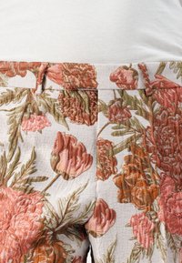 Blomstrete bukser med hevet, teksturert broderi av rosa og oransje blomster på en lys bakgrunn, med en tettsittende midjebånd.