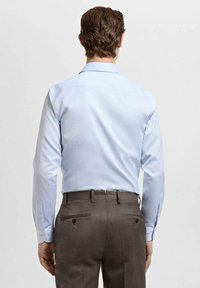 Svetlo modra srajca z dolgimi rokavi, klasičnim ovratnikom, gumbi na manšetah in gladko teksturo, kombinirana z rjavimi hlačami po meri z vidnimi zadnjimi žepi.