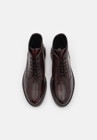 Botas de tobillo de cuero burdeos oscuro con textura suave, punta redonda y cordones negros; presenta detalles de costura y una suela en contraste.