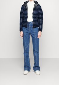 Giacca imbottita blu navy con zip sul davanti e rivestimento grigio, abbinata a jeans azzurri chiari con cuciture uniche e gamba ampia, e sneaker bianche.