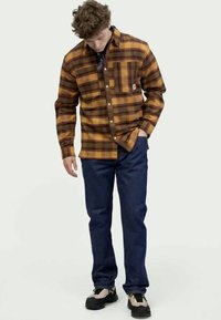 Brown and yellow plaid flannel shirt with a chest pocket, button-up design, paired with dark blue jeans and black sneakers.