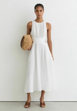 Femeie cu părul împletit într-o rochie midi albă fără mâneci, ținând o geantă clutch împletită și purtând sandale maro cu toc și vârf deschis.