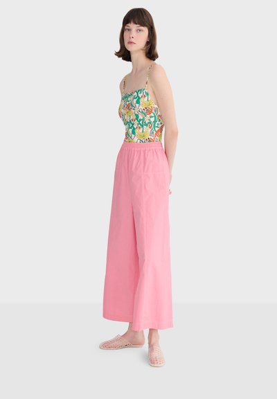 Jeune femme portant un haut à bretelles fleuri, un pantalon large rose taille haute et des chaussures transparentes tissées sans lacets, debout devant un fond uni.