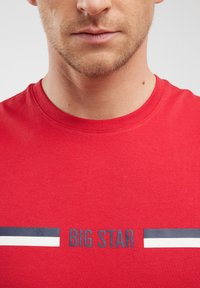 Sarkans T-krekls, kas izgatavots no kokvilnas, ar apaļu izgriezumu un grafisku druku ar tekstu "BIG STAR" tumši zilā krāsā. Ietver svītrotes akcentus.